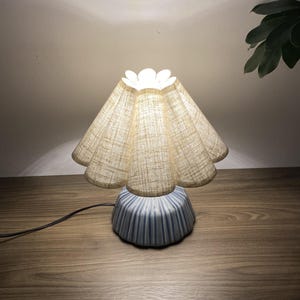 以下が含まれることがあります： ベージュのリネンのシェードと水色のセラミックベースの小さな装飾的なテーブルランプ。シェードはスカラップエッジで、ベースは垂直の溝があります。 ランプは差し込まれており、木製の表面に置かれています。