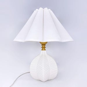 Op de afbeelding: Witte keramische tafellamp met een witte geplooide stoffen lampenkap. De lampvoet heeft een getextureerd bladerenontwerp.