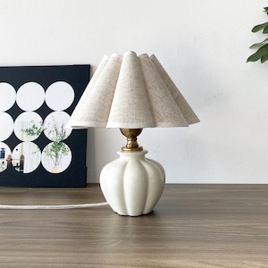 Puede incluir: Una pequeña lámpara de mesa color crema con una pantalla beige plisada. La lámpara tiene una base de cerámica blanca acanalada y un cuello color latón. La lámpara está sobre una superficie de madera.