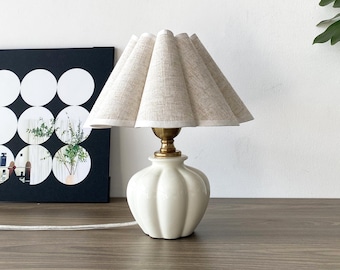 Könnte beinhalten: Eine kleine cremefarbene Tischlampe mit einem plissierten beigefarbenen Lampenschirm. Die Lampe hat einen weißen, gerippten Keramikfuß und einen messingfarbenen Hals. Die Lampe steht auf einer Holzoberfläche.