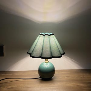 May include: A teal ceramic table lamp with a round base and a pleated lampshade. The lampshade is teal with black trim and a scalloped edge. The lamp is on a wooden surface and has a black cord.