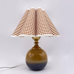 May include: A decorative table lamp with a brown and white gingham shade. The lamp has a brass-colored neck and a round, two-tone base in shades of brown and blue. The lamp is plugged into a wall outlet.