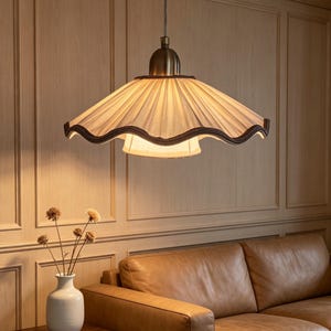 May include: A pendant light fixture featuring a wide, pleated off-white fabric shade with a dark brown scalloped trim. The lamp includes a brass-toned metal cap at the top and a smaller, secondary inner fabric shade suspended beneath the main shade.