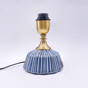 May include: A small, blue ceramic table lamp with a ribbed design. The lamp has a brass-coloured base and a black lamp socket. A black power cord extends from the base. The lamp is designed for indoor use.