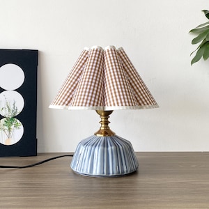 May include: A small table lamp with a blue ceramic base and a brown and white gingham lampshade. The lamp has a brass-coloured neck and a white trim on the lampshade. The lamp is on a wooden surface.