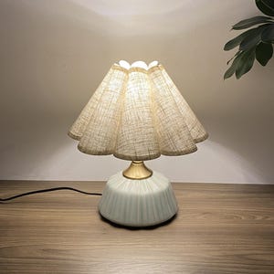 May include: A small, decorative table lamp with a light blue base and a beige, pleated lampshade. The lamp has a gold-colored neck and a black power cord. The lamp is on a wooden surface.