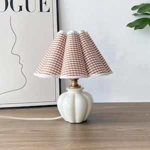 May include: A small table lamp with a white ceramic base and a brown and white gingham lampshade. The lamp has a gold-coloured metal neck and a white cord. The lamp is on a wooden surface.