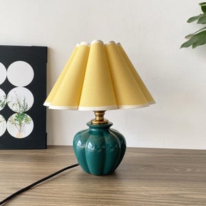 May include: A small table lamp featuring a pleated yellow lampshade and a teal ceramic base. The base has a rounded, ribbed design and a brass-coloured neck. A black power cord extends from the base.