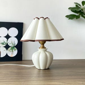 May include: A small, white ceramic table lamp with a pleated off-white shade trimmed with brown. The lamp has a brass base and a ribbed, pumpkin-shaped body. A white cord extends from the base.
