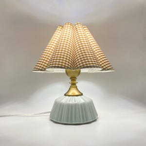 Könnte beinhalten: Dekorative Tischlampe mit hellblauem Keramikfuß und Messingstiel. Der Lampenschirm hat ein plissiertes Design mit einem braun-weißen Vichy-Muster. Die Lampe ist eingeschaltet und beleuchtet den Raum.