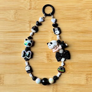 May include: A beaded keychain with a black ring clasp. The keychain features charms of panda bears, hearts, a bow, and a hamburger. The beads are white, black, and iridescent.