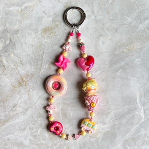May include: A whimsical keychain featuring a variety of charms in shades of pink, yellow, and red. Charms include a donut, a broken heart, a bus, a cupcake, a pinwheel, a strawberry, and a dog. The keychain has a silver ring.