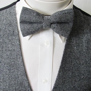 Könnte beinhalten: Eine graue und weiße Fischgrät-Fliege. Die Fliege ist aus einem strukturierten Stoff gefertigt und hat eine klassische Form.