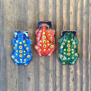 May include: Three colorful frog-shaped magnets. The frogs are painted blue, red, and green, each with yellow and orange polka dots. The word "JAPAN" is printed on the bottom of the red frog. They are set against a wooden background.