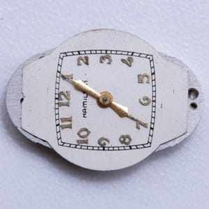 May include: Close-up of a vintage watch face with a white background. The watch face has gold-colored hands and numerals, with the word "HAMILTON" printed on the face. The watch face is surrounded by a silver-colored metal frame.