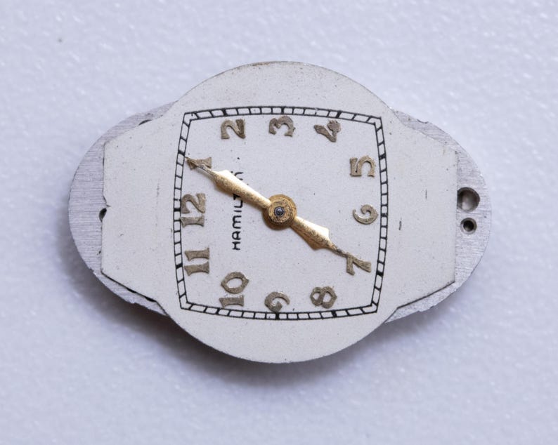 May include: Close-up of a vintage watch face with a white background. The watch face has gold-colored hands and numerals, with the word "HAMILTON" printed on the face. The watch face is surrounded by a silver-colored metal frame.