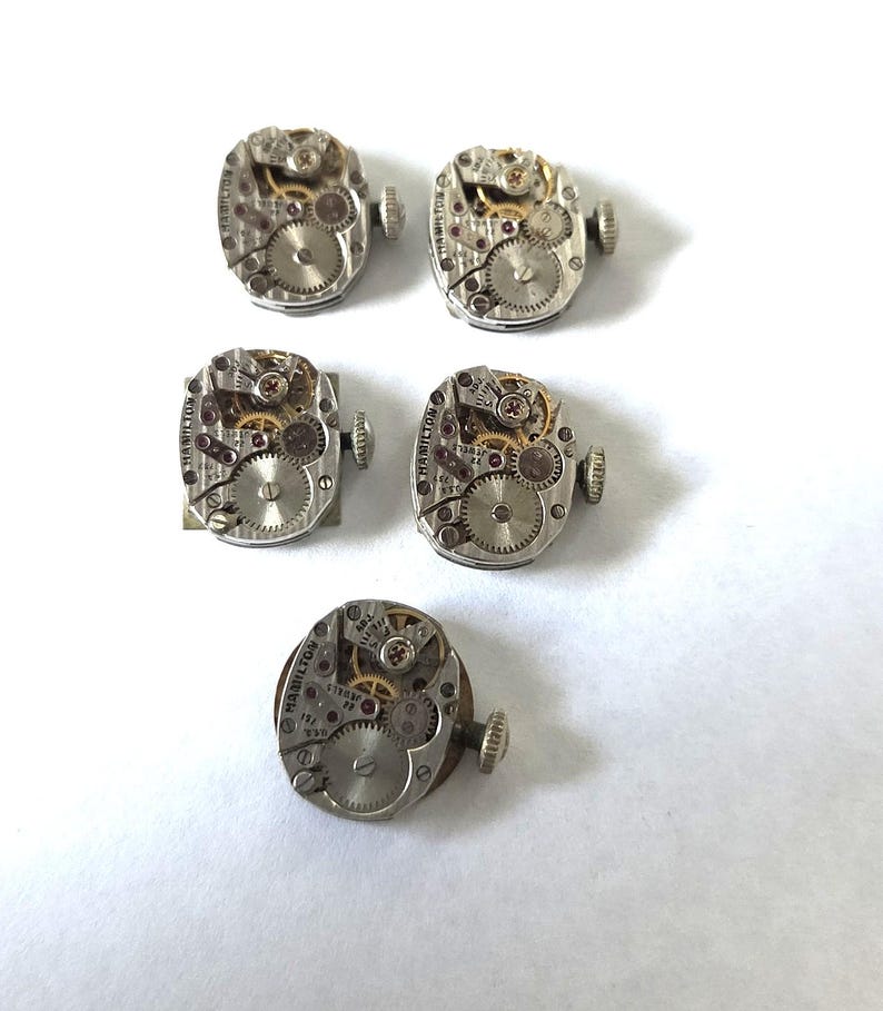 May include: Five vintage Hamilton watch movements. The movements are silver-toned with visible gears, jewels, and other internal components. Each movement has a small crown and is set against a white background. The watch movements are likely for repair or restoration.