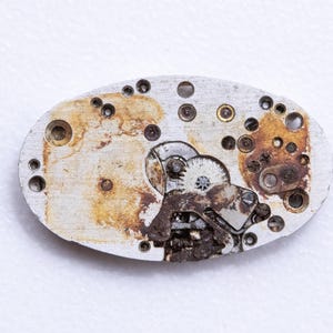 May include: Close-up of a vintage watch movement. The oval-shaped metal plate shows intricate gears, screws, and other components. The metal has a worn, aged appearance with areas of rust and discoloration, suggesting its age and use.