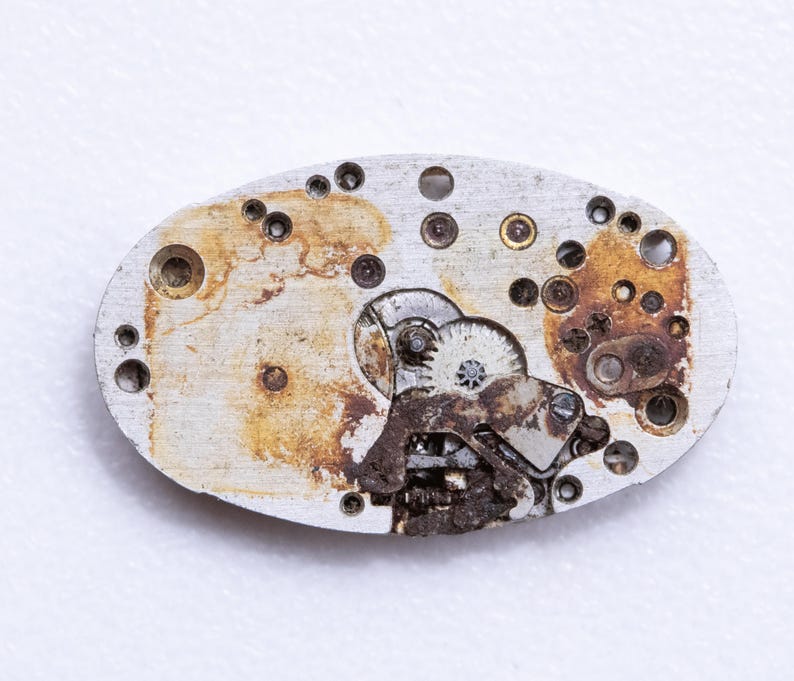 May include: Close-up of a vintage watch movement. The oval-shaped metal plate shows intricate gears, screws, and other components. The metal has a worn, aged appearance with areas of rust and discoloration, suggesting its age and use.