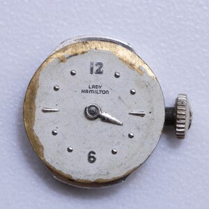 May include: Close-up of a vintage Lady Hamilton watch face. The watch face is white with black numerals at 12 and 6, and silver hands. The watch has a silver case with a gold-toned edge and a silver winding stem.