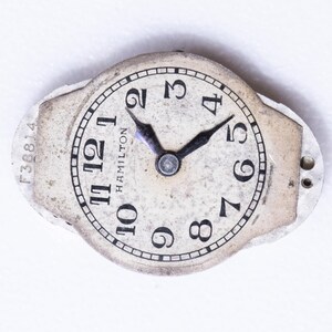 May include: A vintage Hamilton watch face with a cream-colored dial and black numerals. The watch face has black hands and the word "HAMILTON" is printed on the dial. The watch face is surrounded by a silver-colored metal frame. The serial number F38814 is visible.