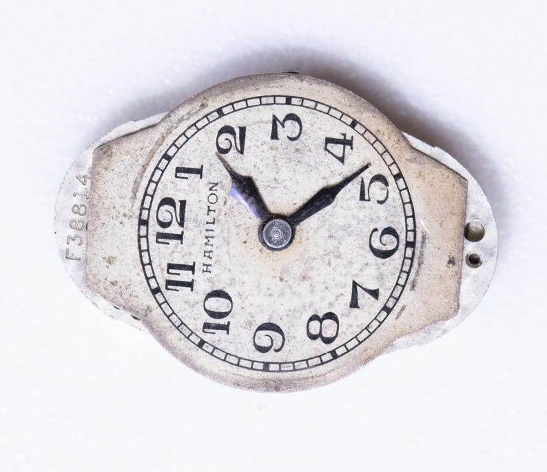 May include: A vintage Hamilton watch face with a cream-colored dial and black numerals. The watch face has black hands and the word "HAMILTON" is printed on the dial. The watch face is surrounded by a silver-colored metal frame. The serial number F38814 is visible.