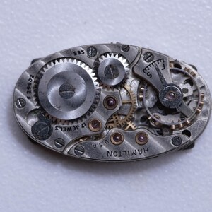 May include: Close-up of a vintage Hamilton watch movement. The oval-shaped mechanism features numerous gears, wheels, and jewels. The text "17 JEWELS" and "U.S.A." are visible, along with the brand name "HAMILTON". The metal components have a silver finish.