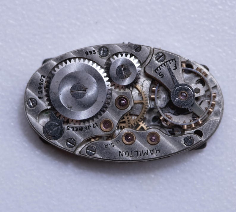 May include: Close-up of a vintage Hamilton watch movement. The oval-shaped mechanism features numerous gears, wheels, and jewels. The text "17 JEWELS" and "U.S.A." are visible, along with the brand name "HAMILTON". The metal components have a silver finish.