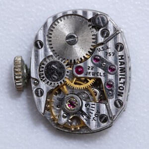 May include: Close-up of a vintage Hamilton watch movement. The silver-toned mechanism features intricate gears, ruby jewels, and the text "HAMILTON" and "U.S.A. 757 22 JEWELS". A small, textured crown is visible on the left side.