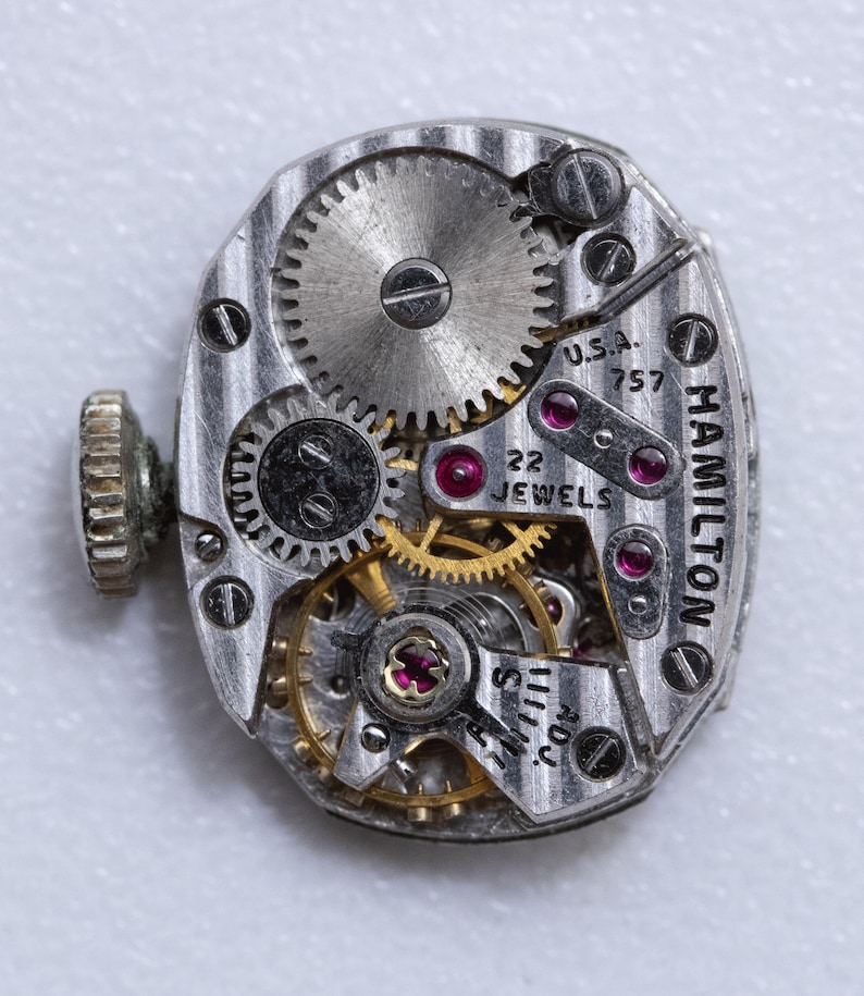 May include: Close-up of a vintage Hamilton watch movement. The silver-toned mechanism features intricate gears, ruby jewels, and the text "HAMILTON" and "U.S.A. 757 22 JEWELS". A small, textured crown is visible on the left side.