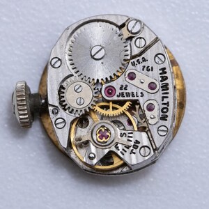 May include: Close-up of a vintage Hamilton watch movement. The mechanism features intricate gears, jewels, and the text "22 JEWELS" and "U.S.A. 761". The watch has a gold-toned outer casing and a silver-colored inner mechanism.
