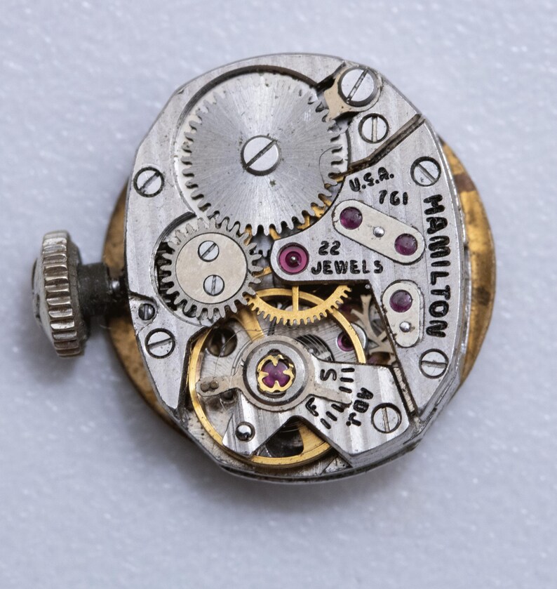 May include: Close-up of a vintage Hamilton watch movement. The mechanism features intricate gears, jewels, and the text "22 JEWELS" and "U.S.A. 761". The watch has a gold-toned outer casing and a silver-colored inner mechanism.