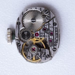 May include: Close-up of a vintage Hamilton watch movement. The silver-toned mechanism features intricate gears, ruby jewels, and the text "22 JEWELS" and "U.S.A. 760". The watch's winding stem is visible on the left.