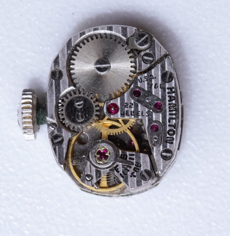 May include: Close-up of a vintage Hamilton watch movement. The silver-toned mechanism features intricate gears, ruby jewels, and the text "22 JEWELS" and "U.S.A. 760". The watch's winding stem is visible on the left.