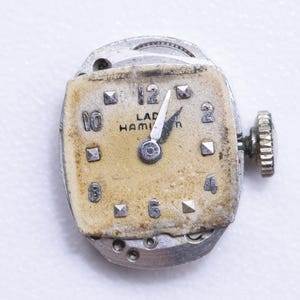 May include: Close-up of a vintage watch movement. The watch face is a faded beige with black numerals and the text "LAD HAMILTON". The hands point to approximately 12:00. The watch has a silver-colored metal casing and a winding stem.