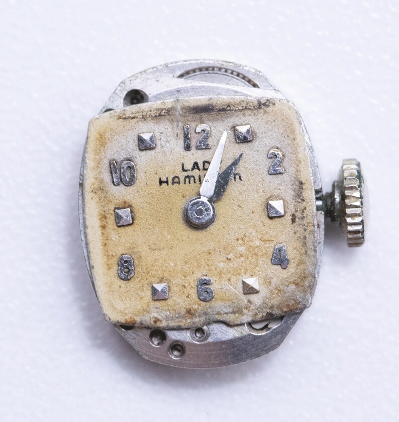 May include: Close-up of a vintage watch movement. The watch face is a faded beige with black numerals and the text "LAD HAMILTON". The hands point to approximately 12:00. The watch has a silver-colored metal casing and a winding stem.
