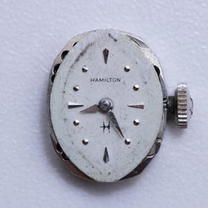 May include: A vintage Hamilton watch face with a silver-tone case and a white dial. The dial features black hands, hour markers, and the brand name "HAMILTON". The watch has a textured crown and a small "H" logo.