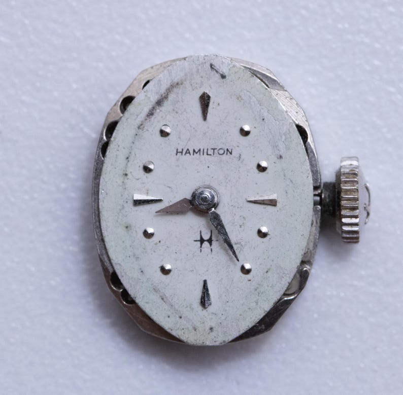 May include: A vintage Hamilton watch face with a silver-tone case and a white dial. The dial features black hands, hour markers, and the brand name "HAMILTON". The watch has a textured crown and a small "H" logo.