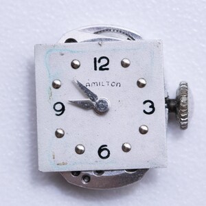 May include: Close-up of a vintage Hamilton watch face. The square, off-white face features black numerals at 12, 3, 6, and 9 o'clock, with silver-tone markers. The word "HAMILTON" is printed above the center. Silver hands point to approximately 9:15. A silver crown is visible.