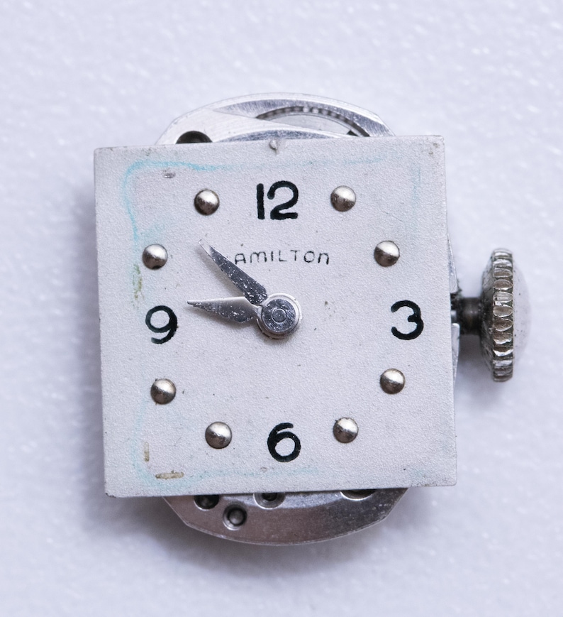 May include: Close-up of a vintage Hamilton watch face. The square, off-white face features black numerals at 12, 3, 6, and 9 o'clock, with silver-tone markers. The word "HAMILTON" is printed above the center. Silver hands point to approximately 9:15. A silver crown is visible.