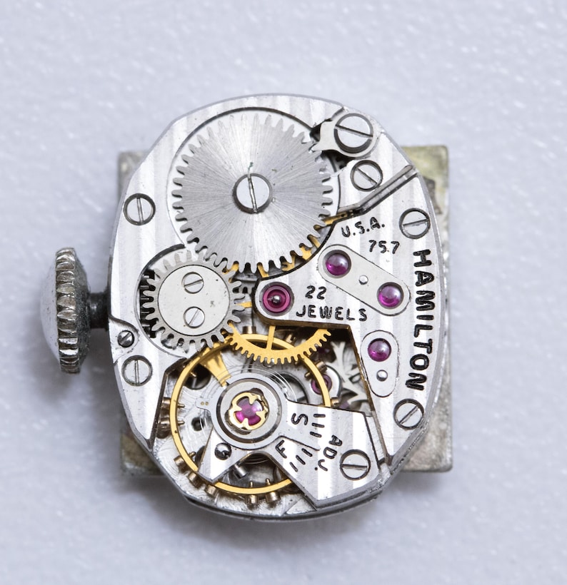 May include: Close-up of a vintage Hamilton watch movement. The silver-toned mechanism features intricate gears, ruby jewels, and the text "U.S.A. 757" and "22 Jewels". The watch has a silver-toned winding stem.
