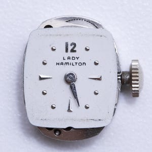 May include: A vintage Lady Hamilton watch face with a white dial. The watch face features the number 12, the brand name, and silver hands. The watch has a silver winding stem and a silver metal casing. The watch is a classic timepiece.