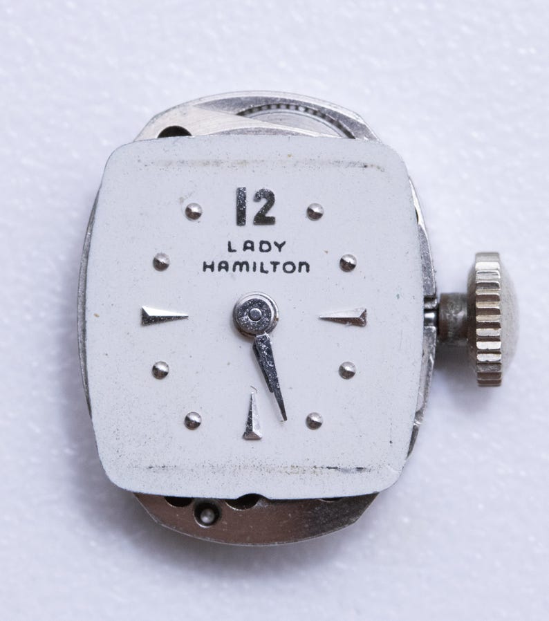 May include: A vintage Lady Hamilton watch face with a white dial. The watch face features the number 12, the brand name, and silver hands. The watch has a silver winding stem and a silver metal casing. The watch is a classic timepiece.