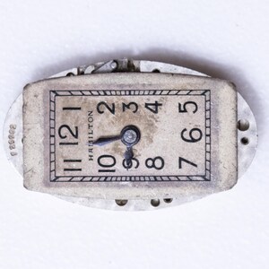 May include: Vintage Hamilton watch face with black numerals on a cream-colored background. The watch face is rectangular with rounded edges and features the brand name "Hamilton". The hands point to approximately 10:09. The watch is set in a silver-colored metal frame.