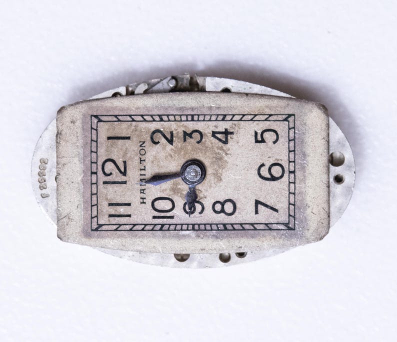 May include: Vintage Hamilton watch face with black numerals on a cream-colored background. The watch face is rectangular with rounded edges and features the brand name "Hamilton". The hands point to approximately 10:09. The watch is set in a silver-colored metal frame.