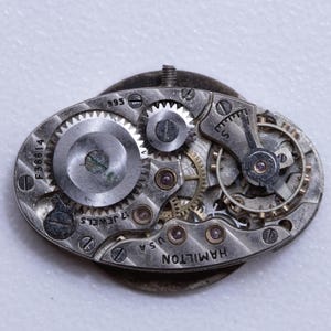 May include: Close-up of a vintage watch movement with intricate gears and components. The silver-toned mechanism features the inscription "HAMILTON U.S.A." and "17 JEWELS." The oval-shaped piece has a small screw and a series of tiny screws holding the parts together.