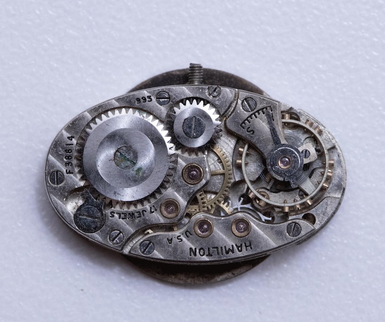 May include: Close-up of a vintage watch movement with intricate gears and components. The silver-toned mechanism features the inscription "HAMILTON U.S.A." and "17 JEWELS." The oval-shaped piece has a small screw and a series of tiny screws holding the parts together.
