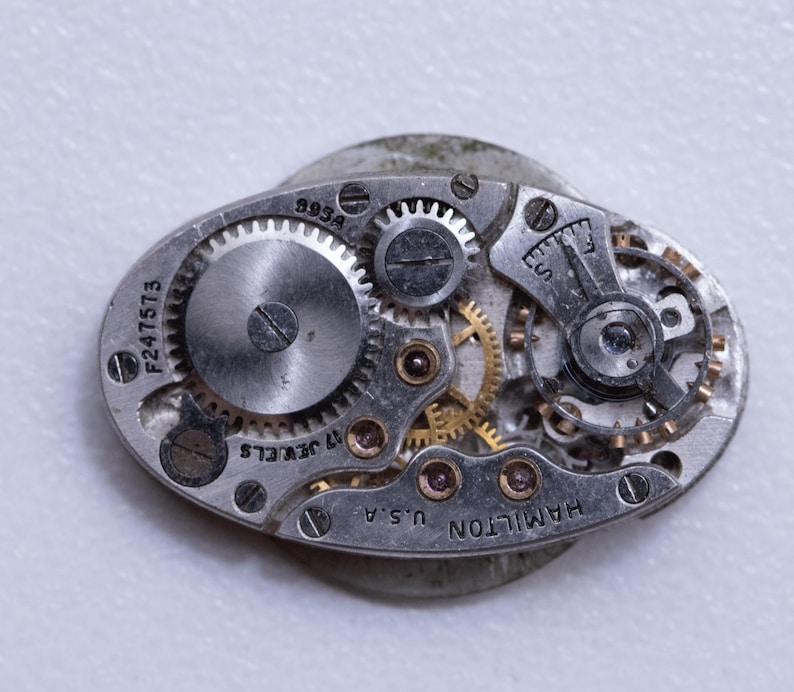 May include: Close-up of a vintage watch movement with visible gears, screws, and other mechanical components. The watch mechanism is oval-shaped and features the text "HAMILTON U.S.A." and "17 JEWELS". The serial number "F247573" is also visible.