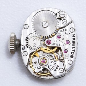 May include: Close-up of a vintage Hamilton watch movement. The silver-toned mechanism features intricate gears, ruby jewels, and the text "U.S.A. 757" and "22 JEWELS". A winding stem is visible on the left side.