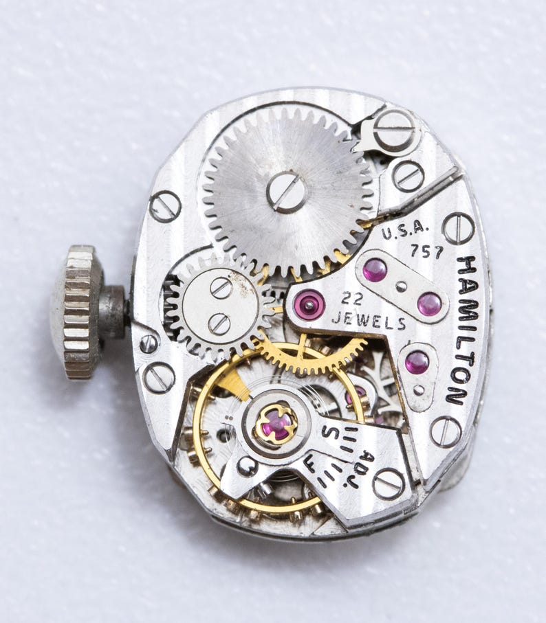 May include: Close-up of a vintage Hamilton watch movement. The silver-toned mechanism features intricate gears, ruby jewels, and the text "U.S.A. 757" and "22 JEWELS". A winding stem is visible on the left side.
